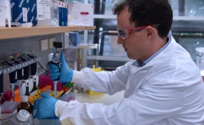 A man in a white lab coat, goggles and gloves in profile, working at a laboratory bench.